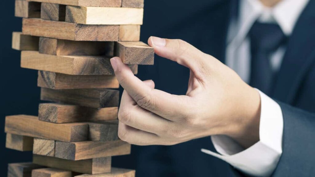 man in business suit pulls a piece out of wobbly wooden tower